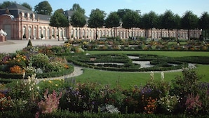 Un jardín formal con césped cuidado, parterres de flores y un edificio clásico con columnas.