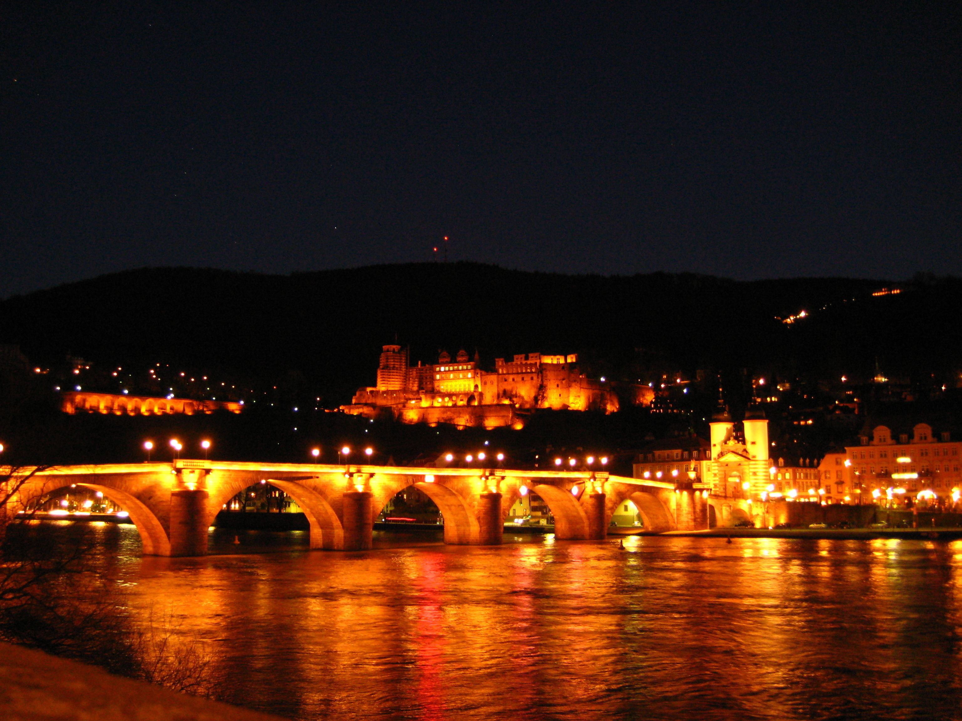 A nighttime view of a bridge with illuminated arches over a river, leading to a historic castle with multiple towers and walls.