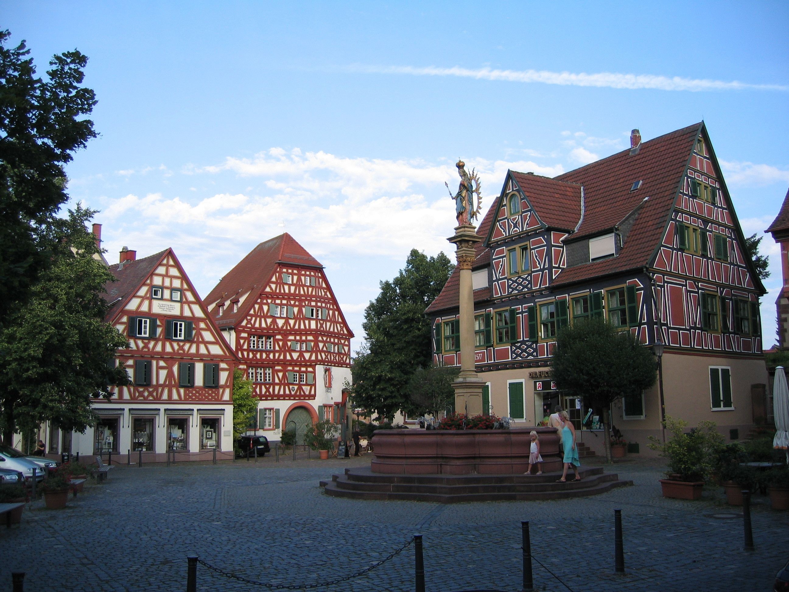 A traditional European town square with historic half-timbered buildings, a central fountain, and cobblestone pavement.