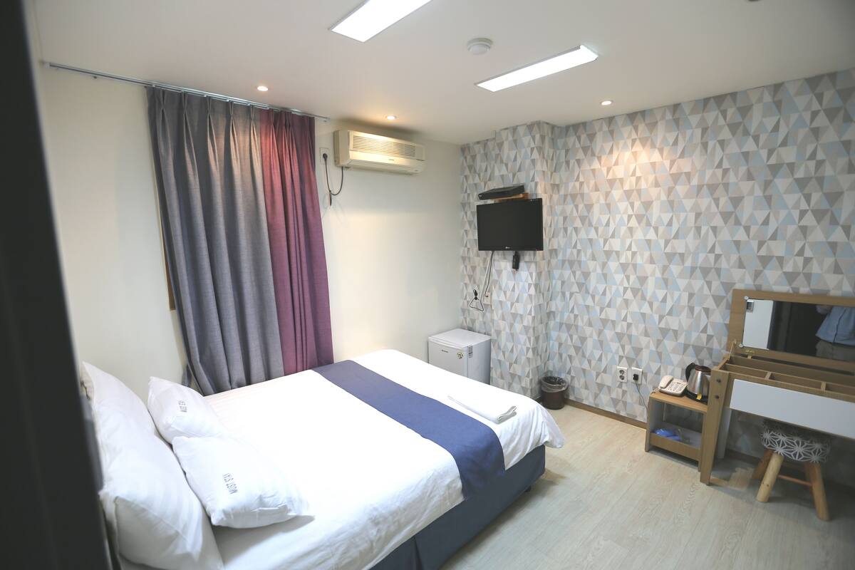 A hotel room with a bed, a desk with a chair, a television mounted on the wall, and an air conditioning unit.