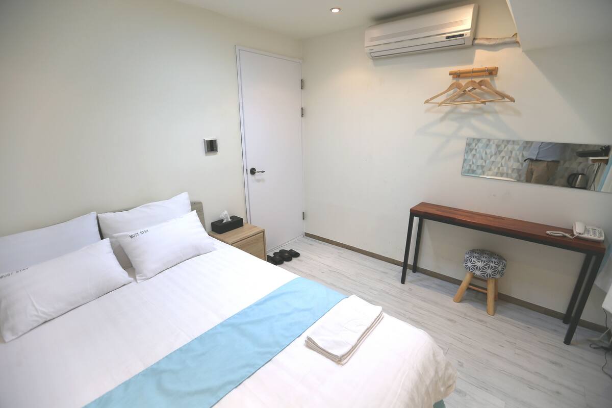 A neatly made bed with pillows, a bedside table with a lamp, a desk with a chair, and a hanging clothes rack.