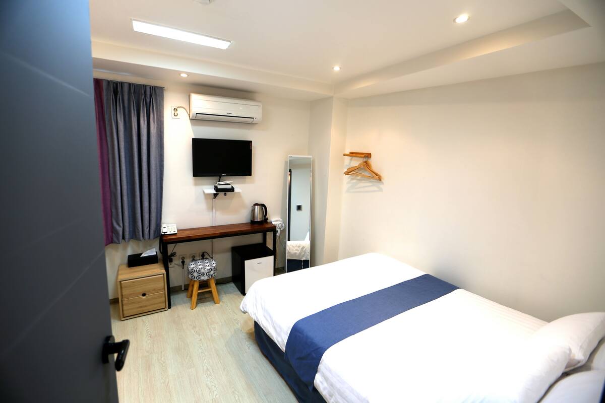 A hotel room with a bed, a desk with a chair, a wardrobe, a mirror, and an air conditioning unit.
