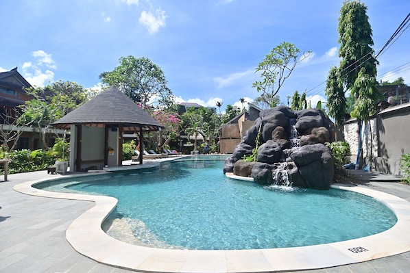 A swimming pool with a waterfall feature, surrounded by lounge chairs and a gazebo. - Puri Cendana Resort Bali (Seminyak)