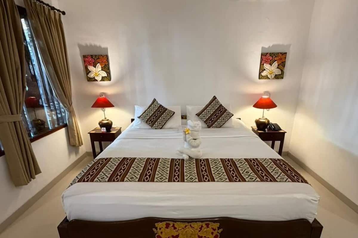 A hotel room with a large bed, two bedside tables, a wall-mounted air conditioner, and a window with curtains.