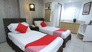 Quarto de hotel com duas camas, cada uma com roupa de cama vermelha e branca, mesa de cabeceira com telefone, ar condicionado fixado na parede, uma pequena planta e uma escrivaninha branca com chaleira.