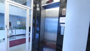Um elevador com visor digital, um botão de alarme e uma placa indicando a capacidade máxima de pessoas.