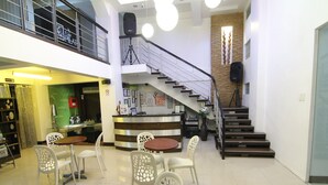 Um hall amplo com um balcão de café, área de estar com mesas e cadeiras, uma escadaria e um balcão de recepção.