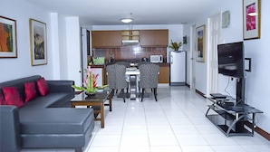 A modern living room with a grey sofa, a dining table with chairs, a kitchen area with cabinets and appliances, and a TV on a stand. - Newgrange Condotel (Quezon City)