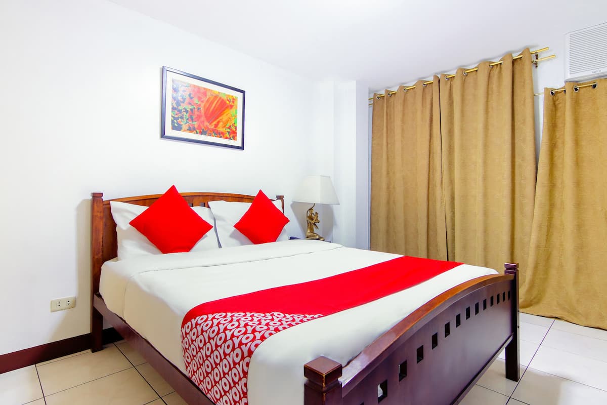 A neatly made bed with a red and white bedspread, a wooden headboard, and a red pillow. There is a framed picture on the wall above the bed, and a window with beige curtains to the right.