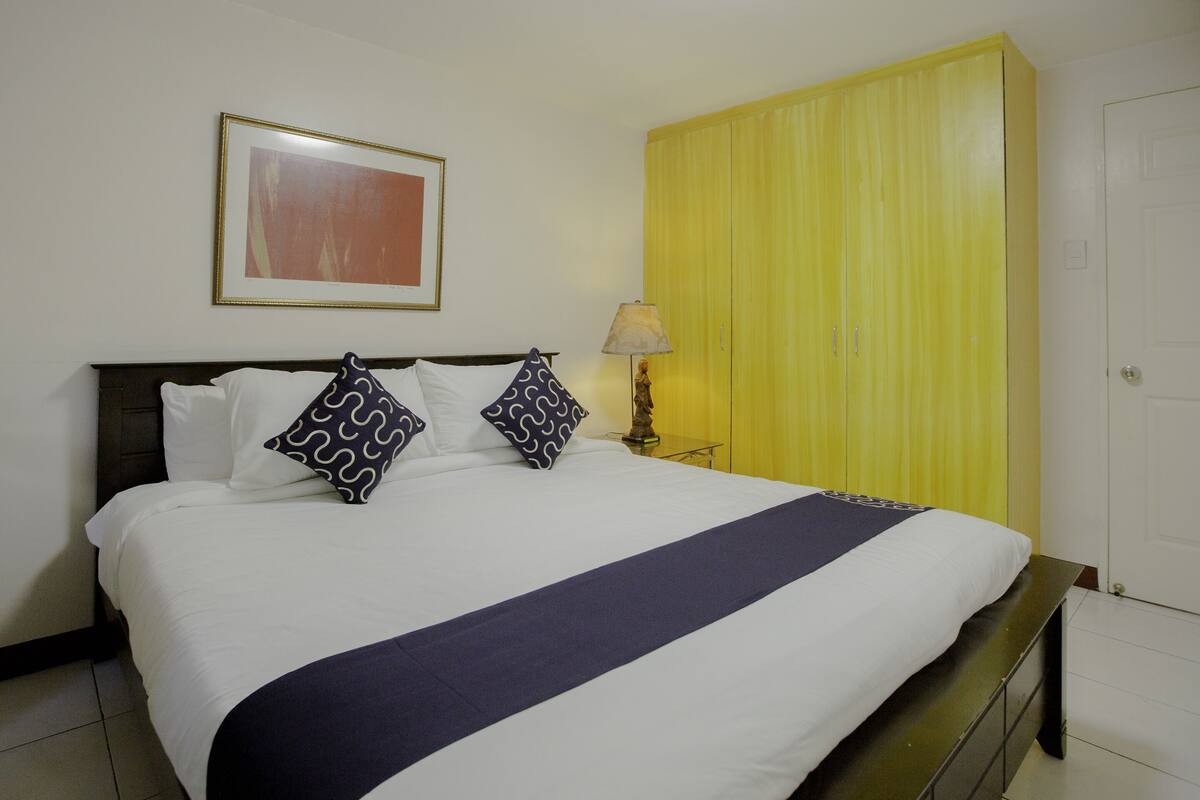 A neatly made bed with a white and navy blue bedspread, two patterned pillows, a framed picture on the wall, a yellow wardrobe, and a bedside lamp.