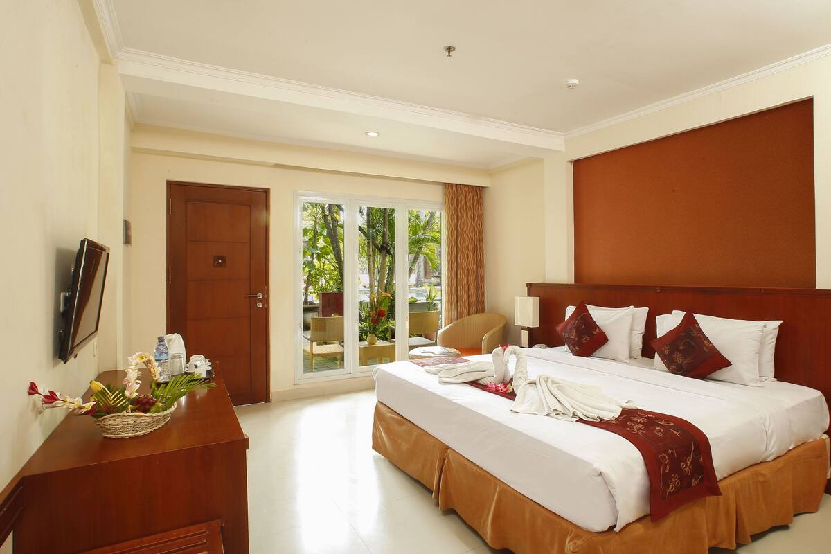 A hotel room with a large bed, a wooden desk, a television, and a balcony with a view of greenery.