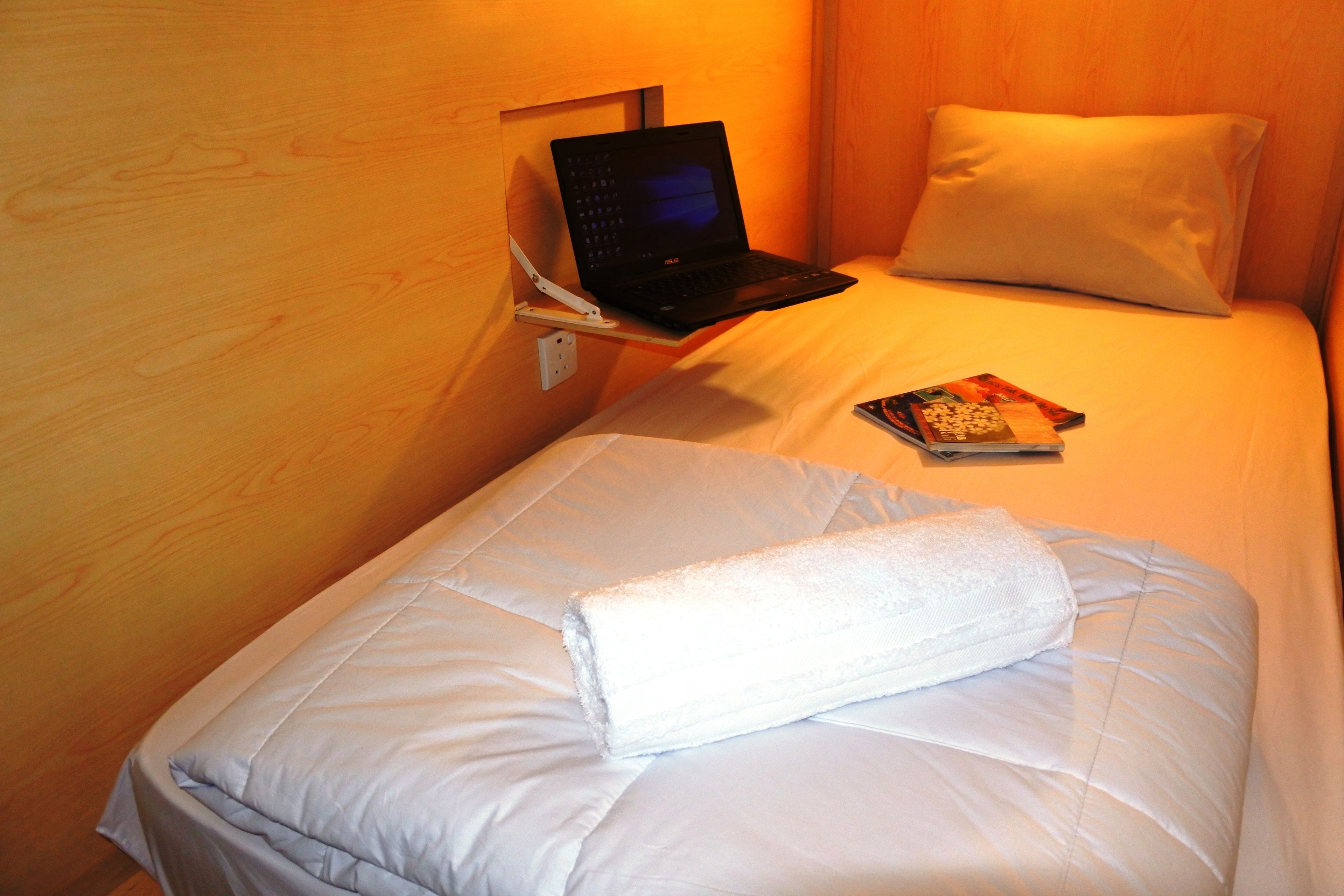 A neatly made bed with a laptop on a bedside table, a rolled-up towel, and a book on the bed.