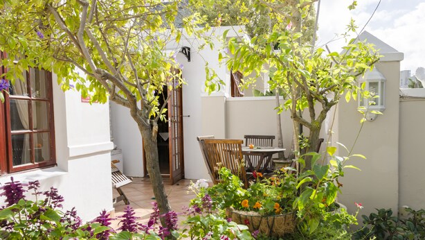 A small, enclosed garden with a wooden table and chairs, surrounded by various plants and flowers. - Brighton Lodge (Gqeberha (Port Elizabeth))