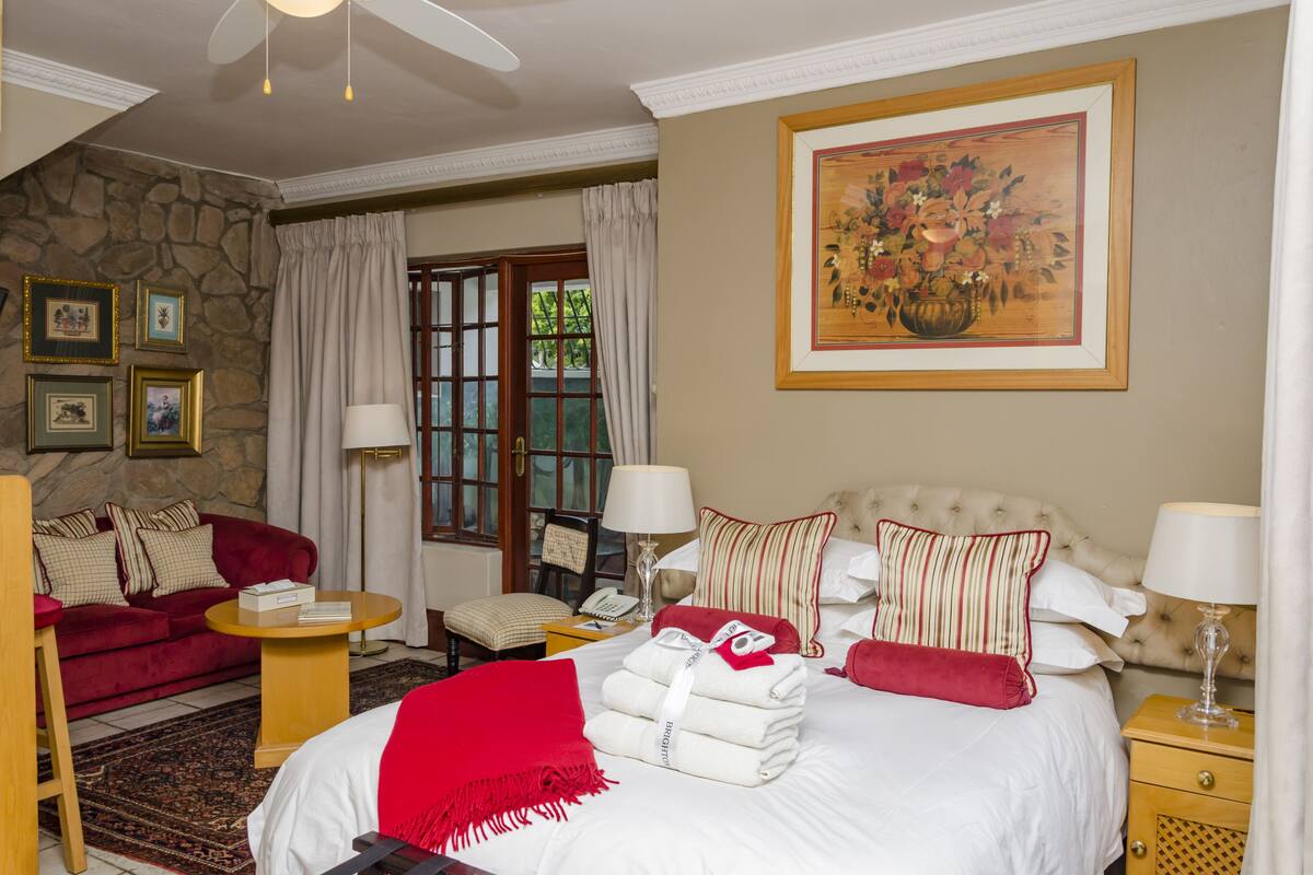 Habitación de hotel con cama, sofá rojo, una mesita, una silla, una mesita de noche y un cuadro en la pared.