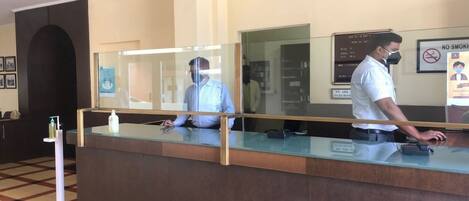 A reception desk with a glass partition, a man wearing a mask, and a 'No Smoking' sign on the wall.