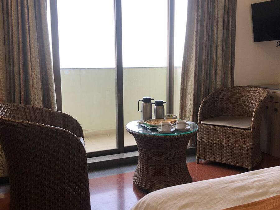 A hotel room with a large sliding glass door, a bed, two wicker chairs, a small round table with a coffee pot and cups, and a TV mounted on the wall.