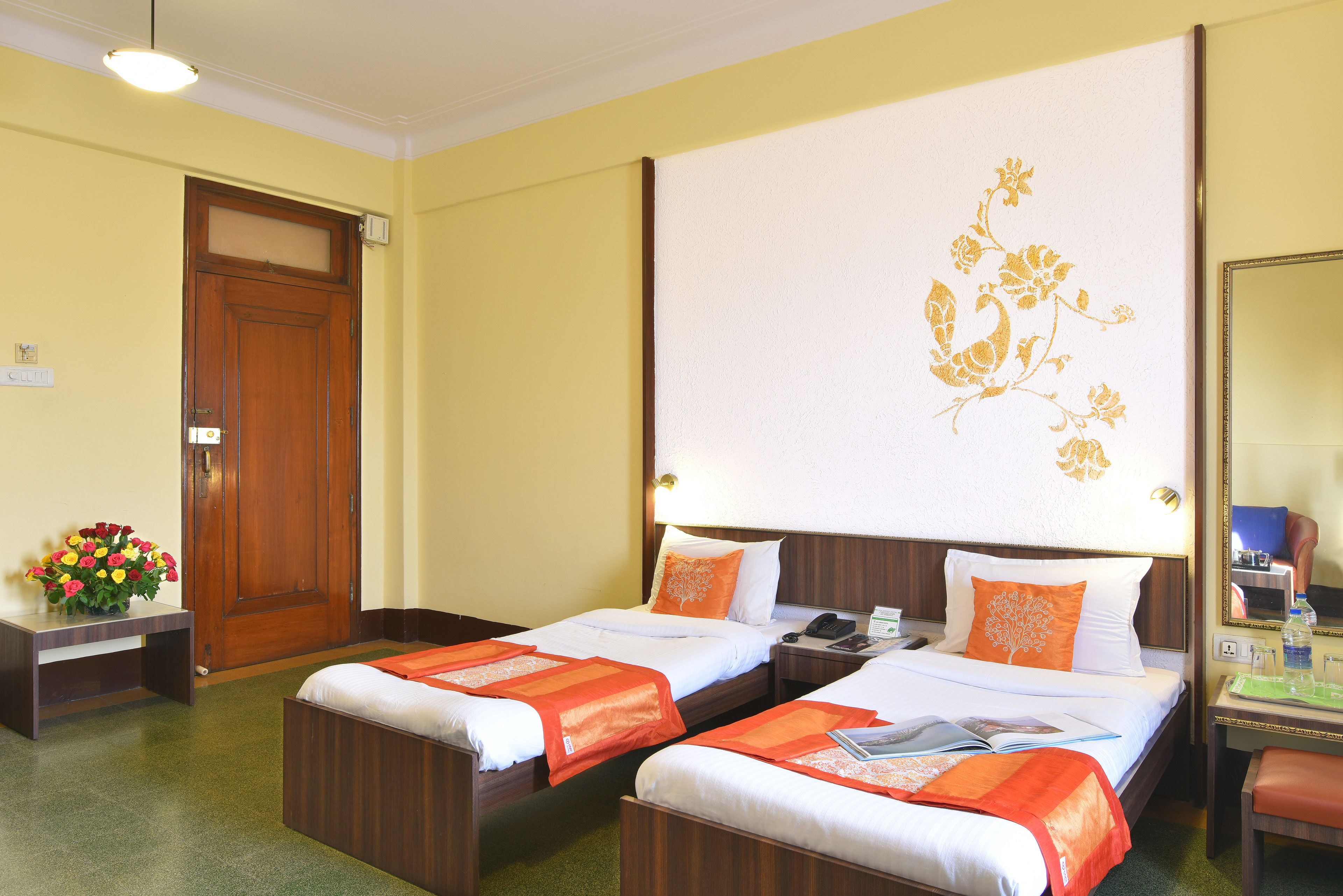 Een hotelkamer met twee eenpersoonsbedden, een houten bureau, een stoel, een nachtkastje, een bedtafel, een lamp, een spiegel en een bloemendecoratie aan de muur.