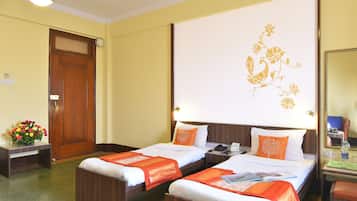 Een hotelkamer met twee eenpersoonsbedden, een houten bureau, een stoel, een nachtkastje, een bedtafel, een lamp, een spiegel en een bloemendecoratie aan de muur.