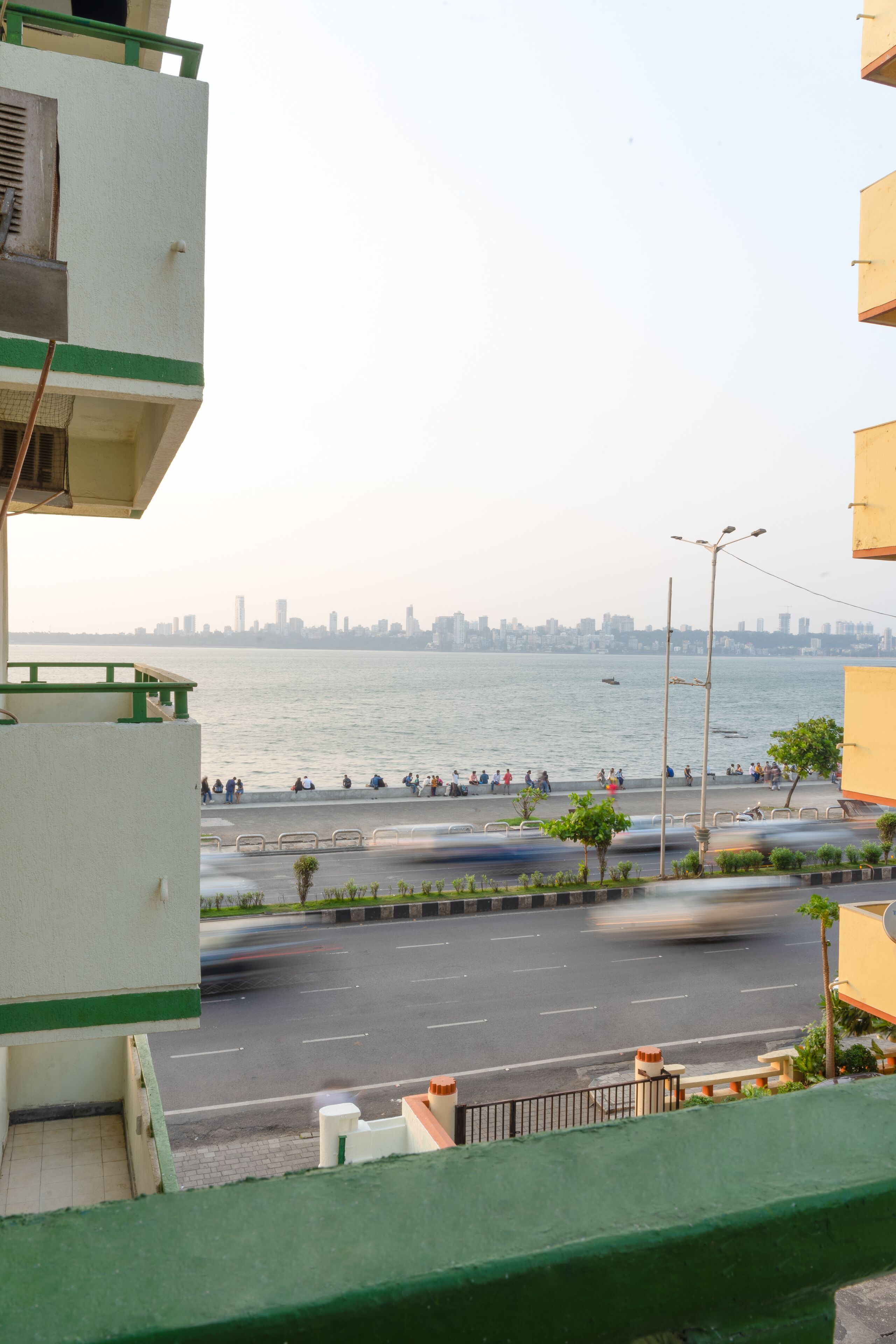 A waterfront view with a city skyline in the background, a road with moving vehicles, and a pedestrian walkway with people.