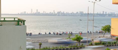 Blick auf das Wasser mit Stadtansicht im Hintergrund, eine Straße mit fahrenden Fahrzeugen und ein Fußweg mit Personen.