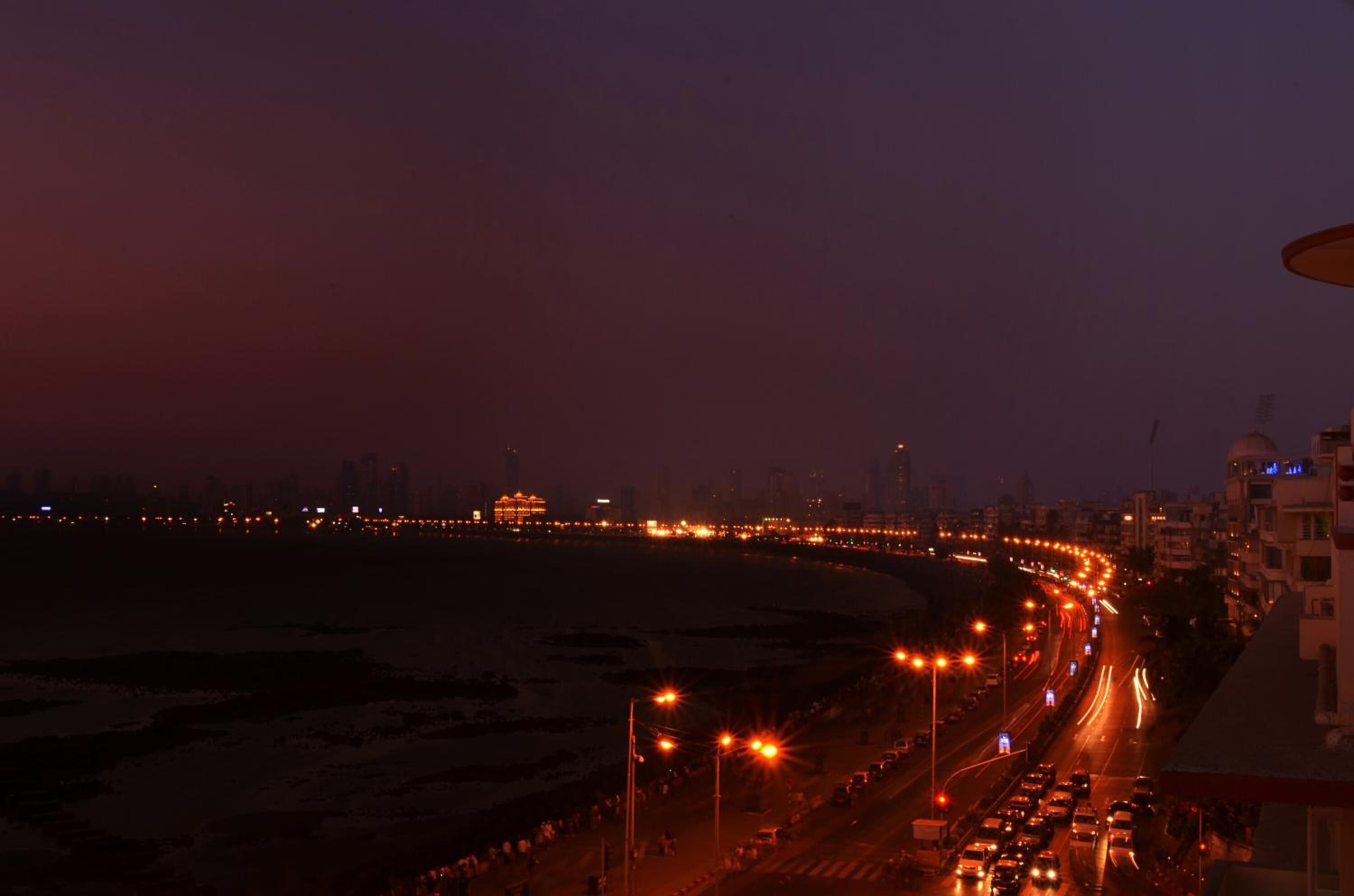 A nighttime cityscape with a curved road leading to a beach, illuminated by streetlights and vehicle headlights.