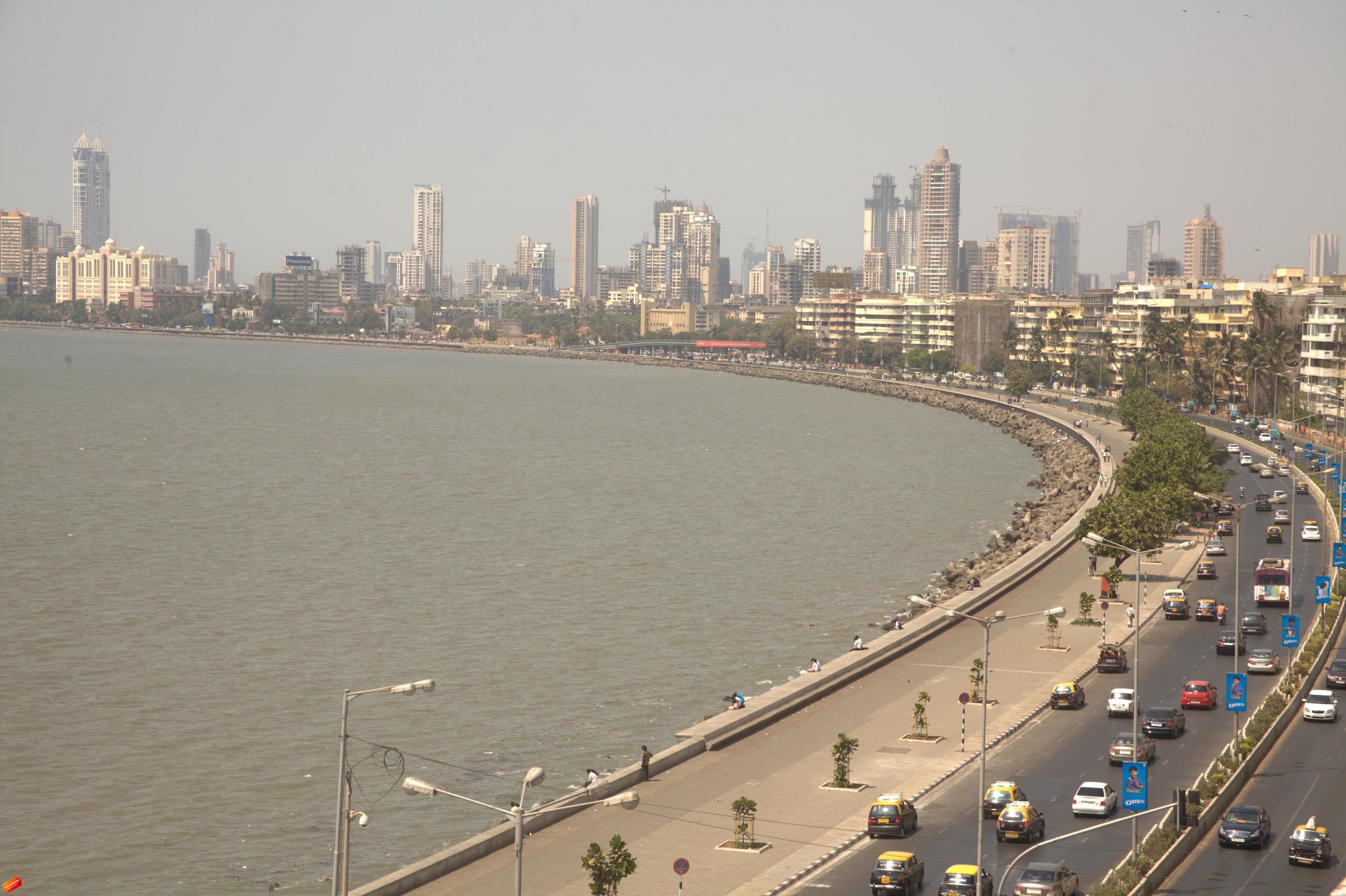 A coastal cityscape with a wide road parallel to the water, lined with buildings and a clear sky.