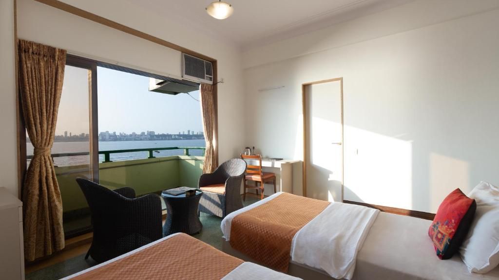 Habitación de hotel con dos camas, un escritorio, una silla y vista a un horizonte urbano a través de amplias ventanas.
