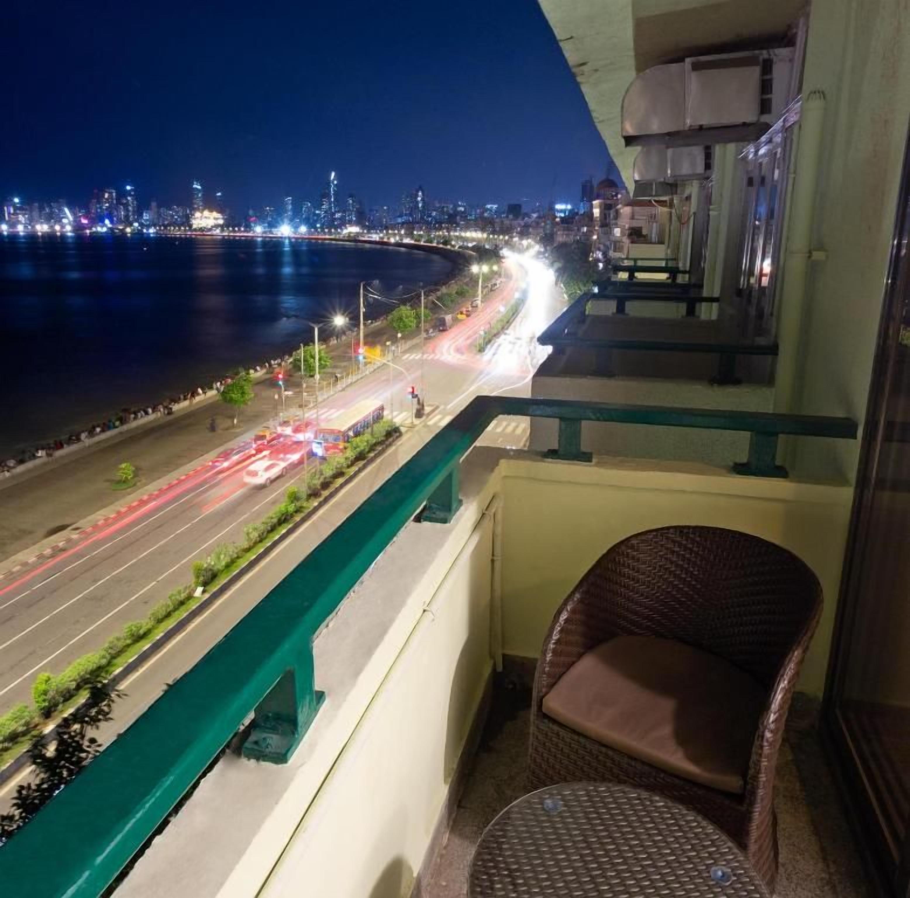 A balcony with a view of a cityscape at night, a wicker chair, and a small table.