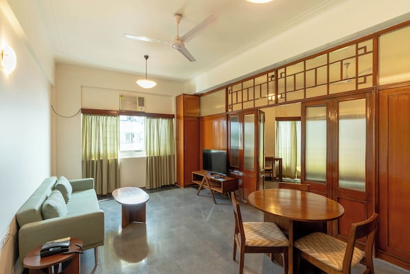 A living room with a sofa, a TV on a wooden stand, a dining table with chairs, and a ceiling fan.