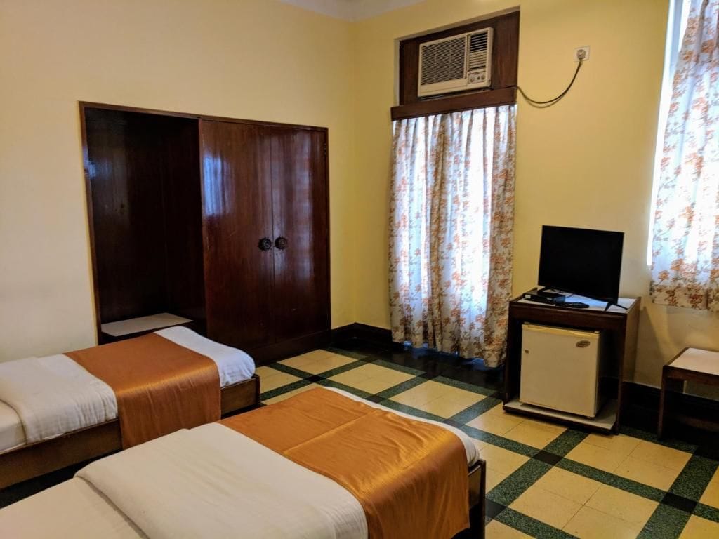 A hotel room with two beds, a television, a small refrigerator, and a window with curtains.