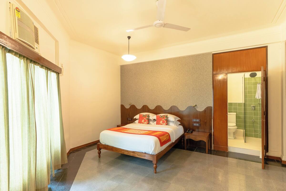 Una habitación de hotel con cama, ventilador de techo, mesita de noche y baño con ducha e inodoro.