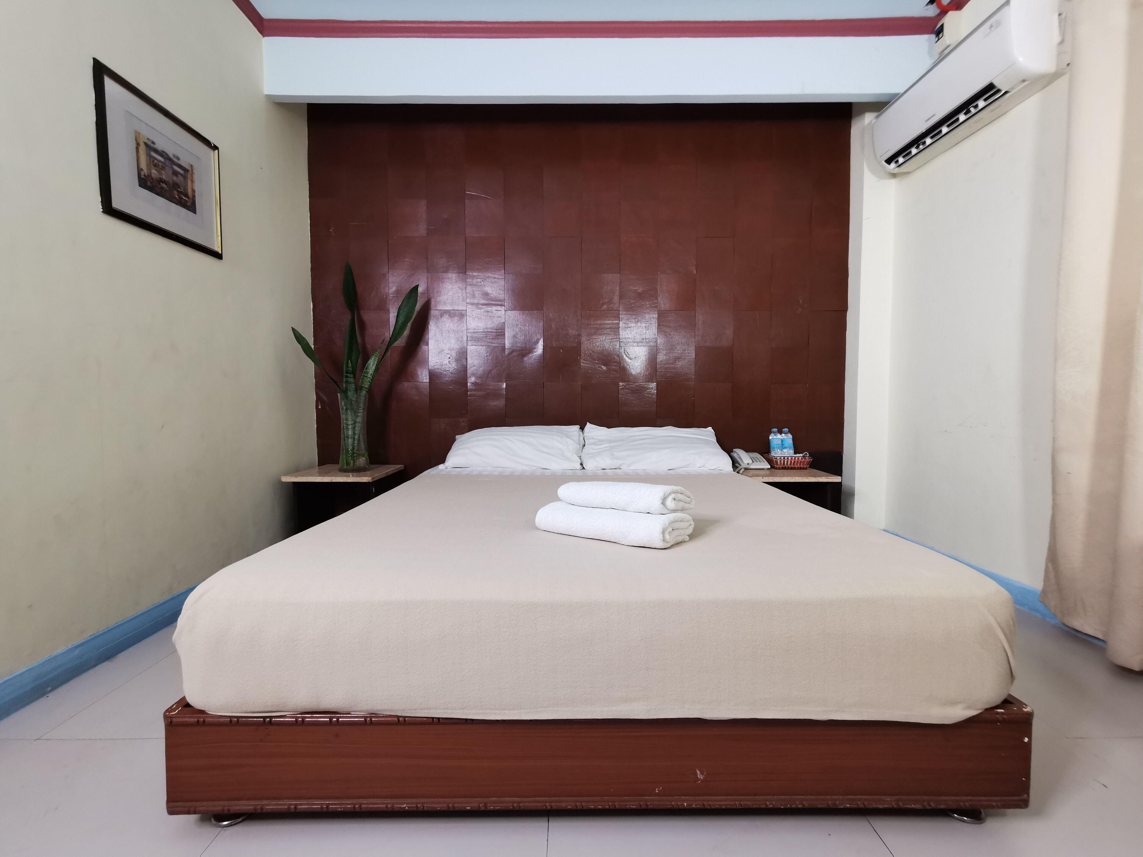A neatly made bed with white linens and towels, a wooden headboard, a small bedside table with a phone and water bottles, and a potted plant.