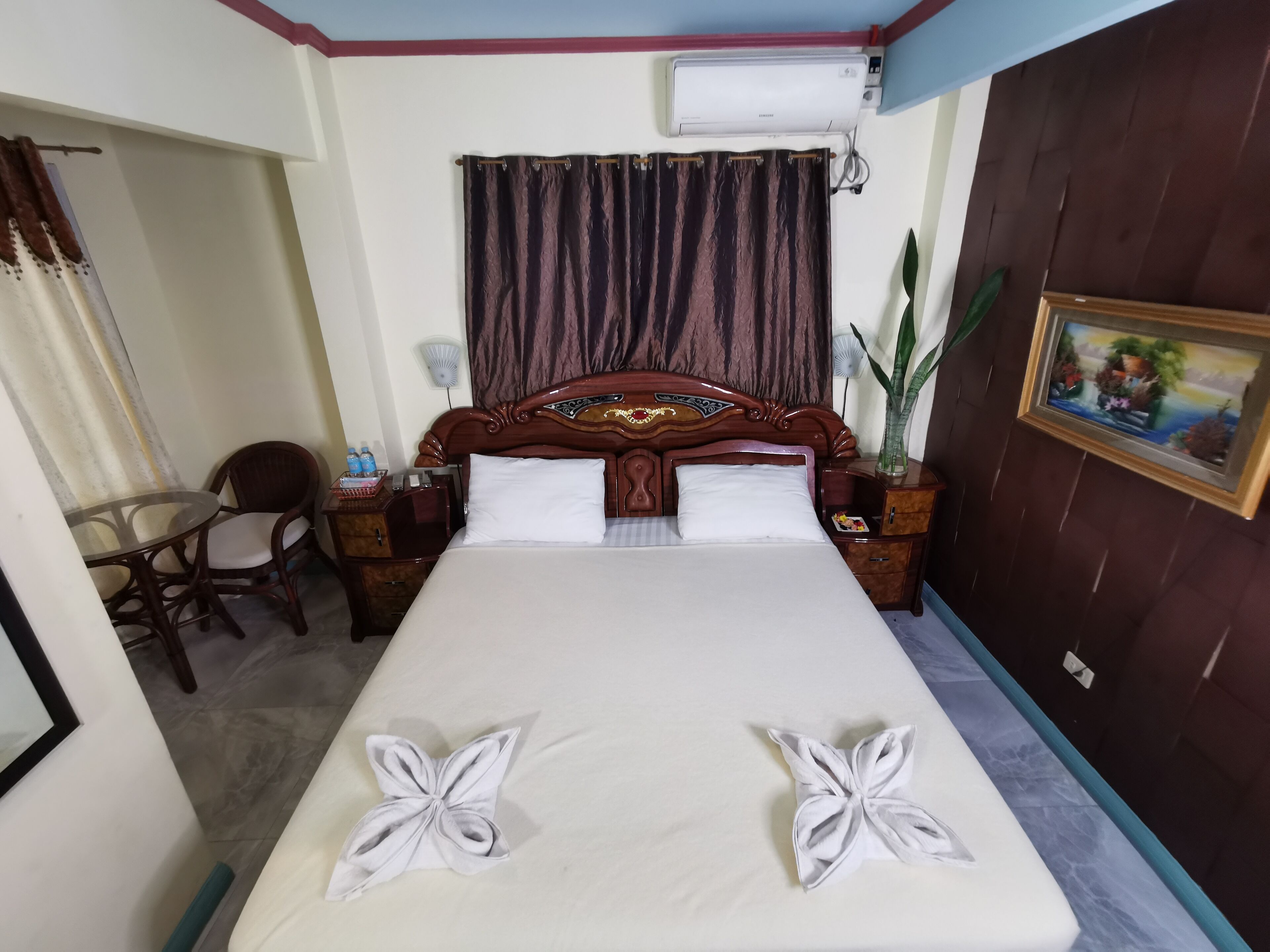 Habitación de hotel con una cama grande, cabecera de madera, mesita de noche, sillón, pequeña mesa y ventana con cortinas.