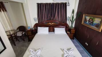 Habitación de hotel con una cama grande, cabecera de madera, mesita de noche, sillón, pequeña mesa y ventana con cortinas.
