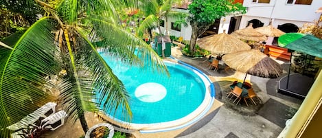 A circular pool surrounded by palm trees and thatched umbrellas, with lounge chairs and a building in the background.