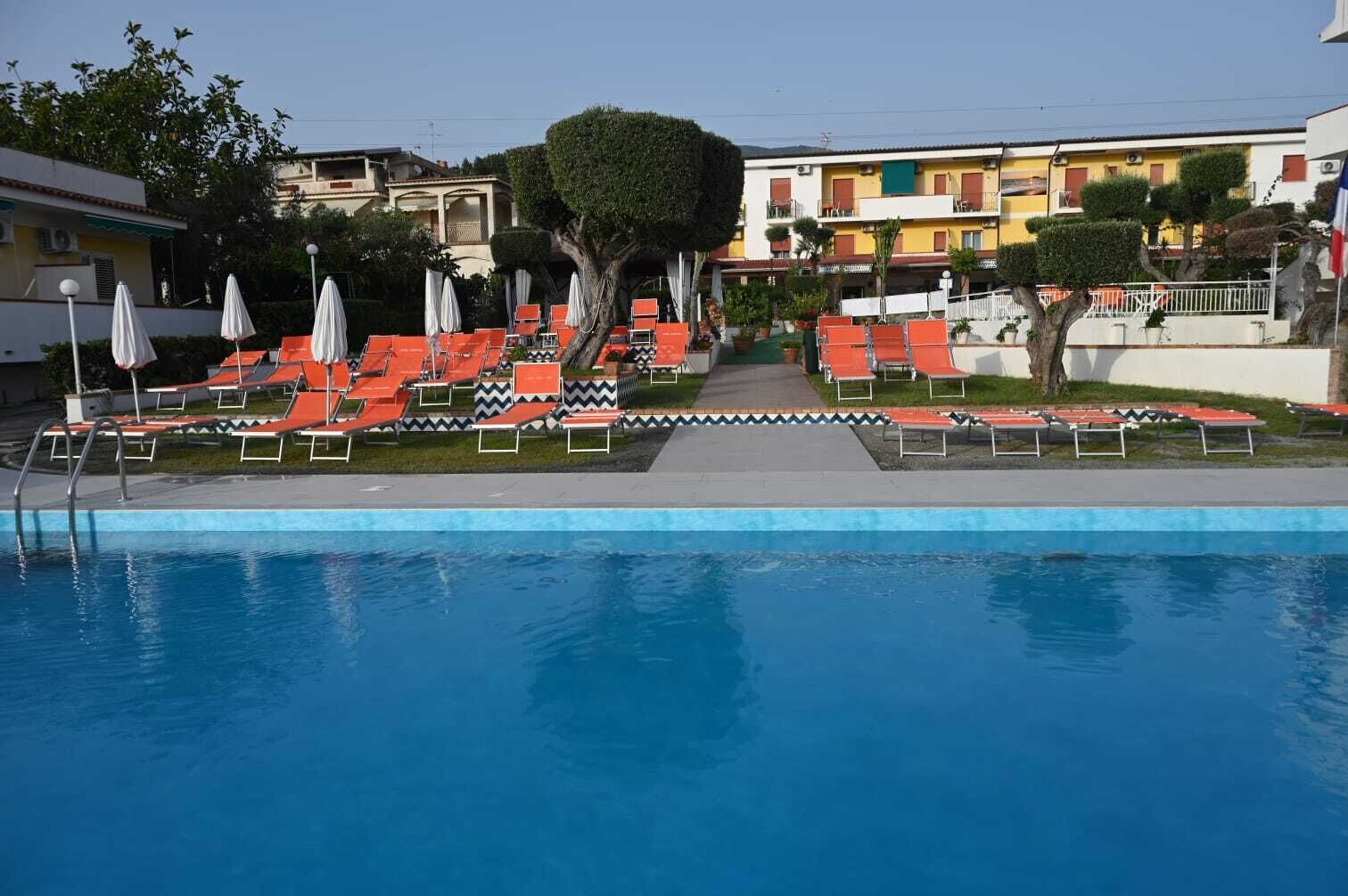 A swimming pool with red lounge chairs and umbrellas, surrounded by a pathway and buildings in the background.