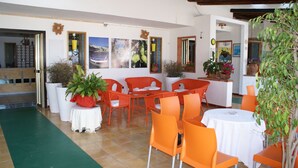 Un área de comedor con sillas y mesas naranja, una alfombra verde y varias plantas en macetas.