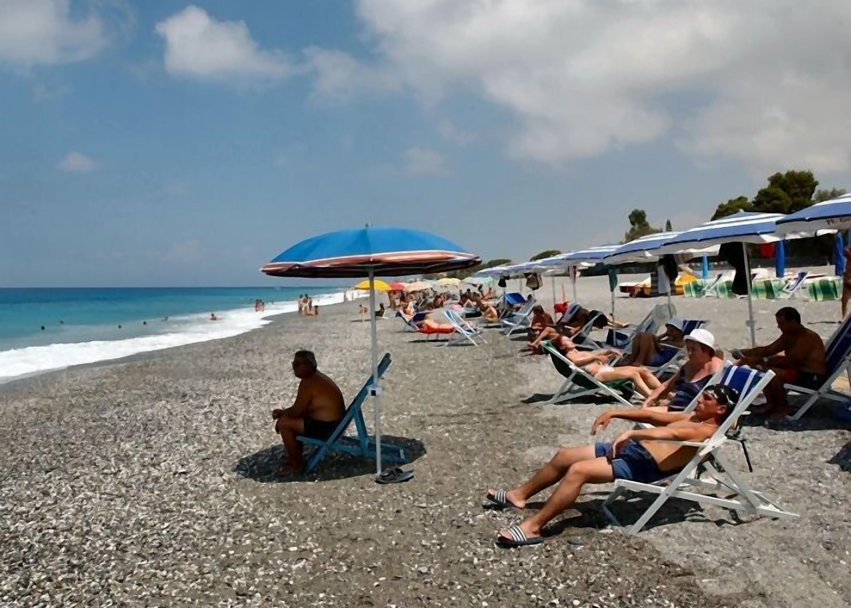 Una playa con gente tomando el sol bajo sombrillas, una costa rocosa y el océano al fondo.