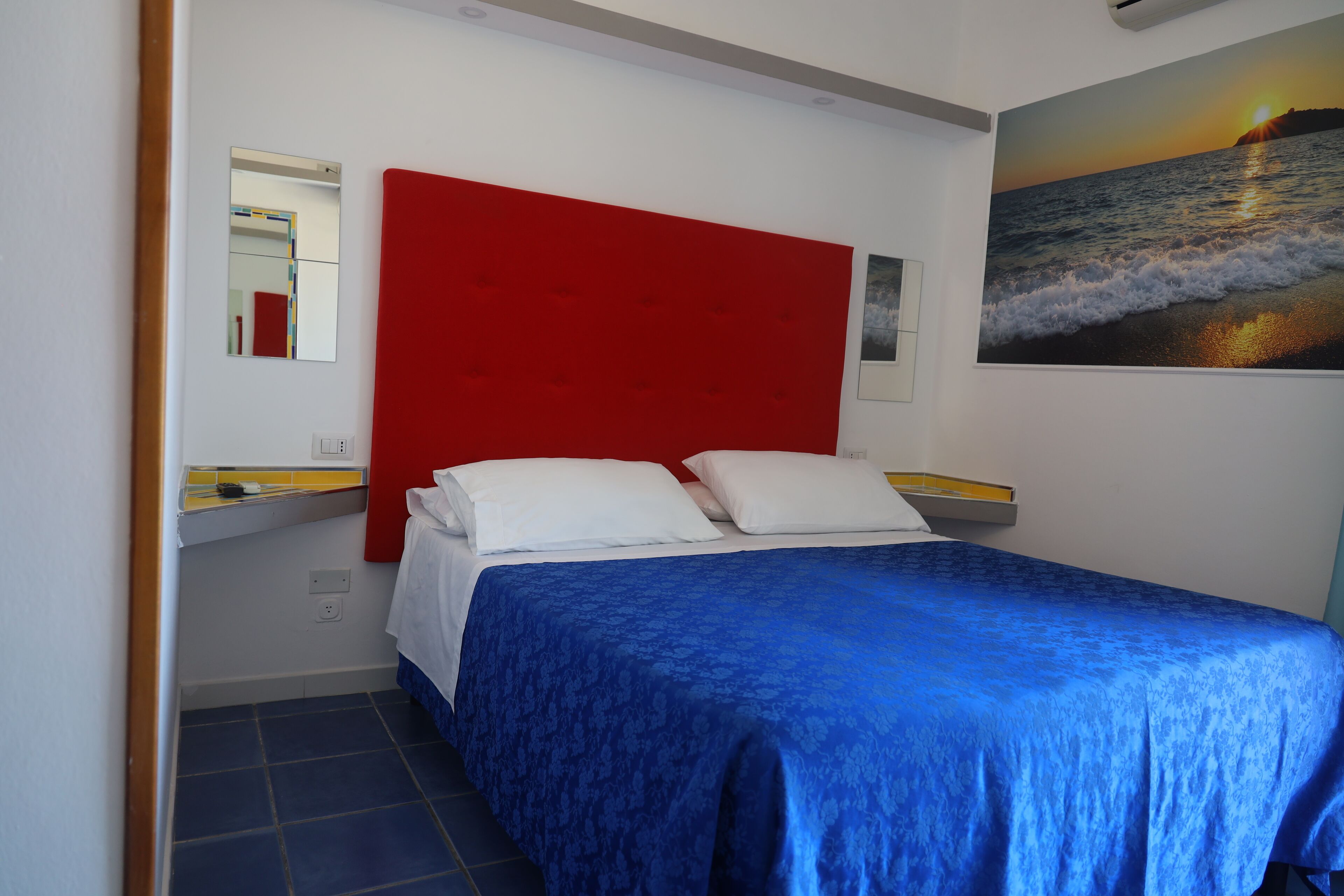 Una camera da letto con una testiera rossa, una coperta blu e un quadro raffigurante un tramonto sull’oceano.