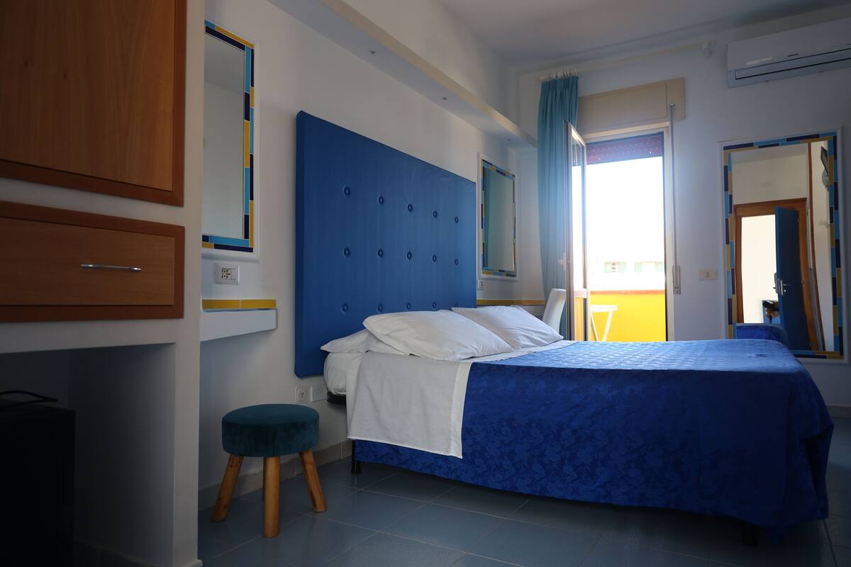 Ein Schlafzimmer mit blauem Kopfteil, einem Bett mit weißer Bettwäsche, einem Holzschrank und einem kleinen Hocker.