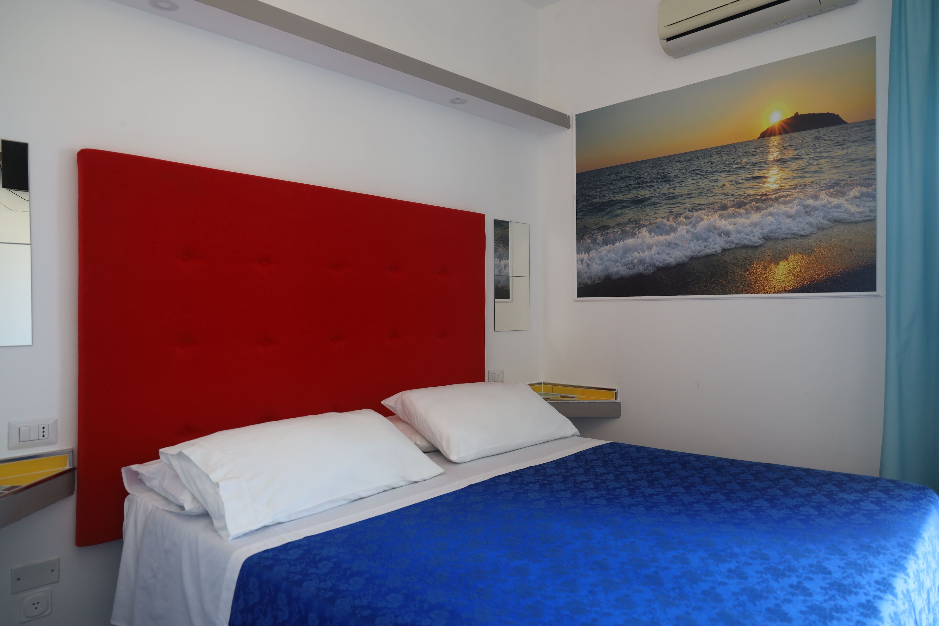 A bedroom with a red headboard, a blue bedspread, white pillows, and a wall-mounted air conditioner.
