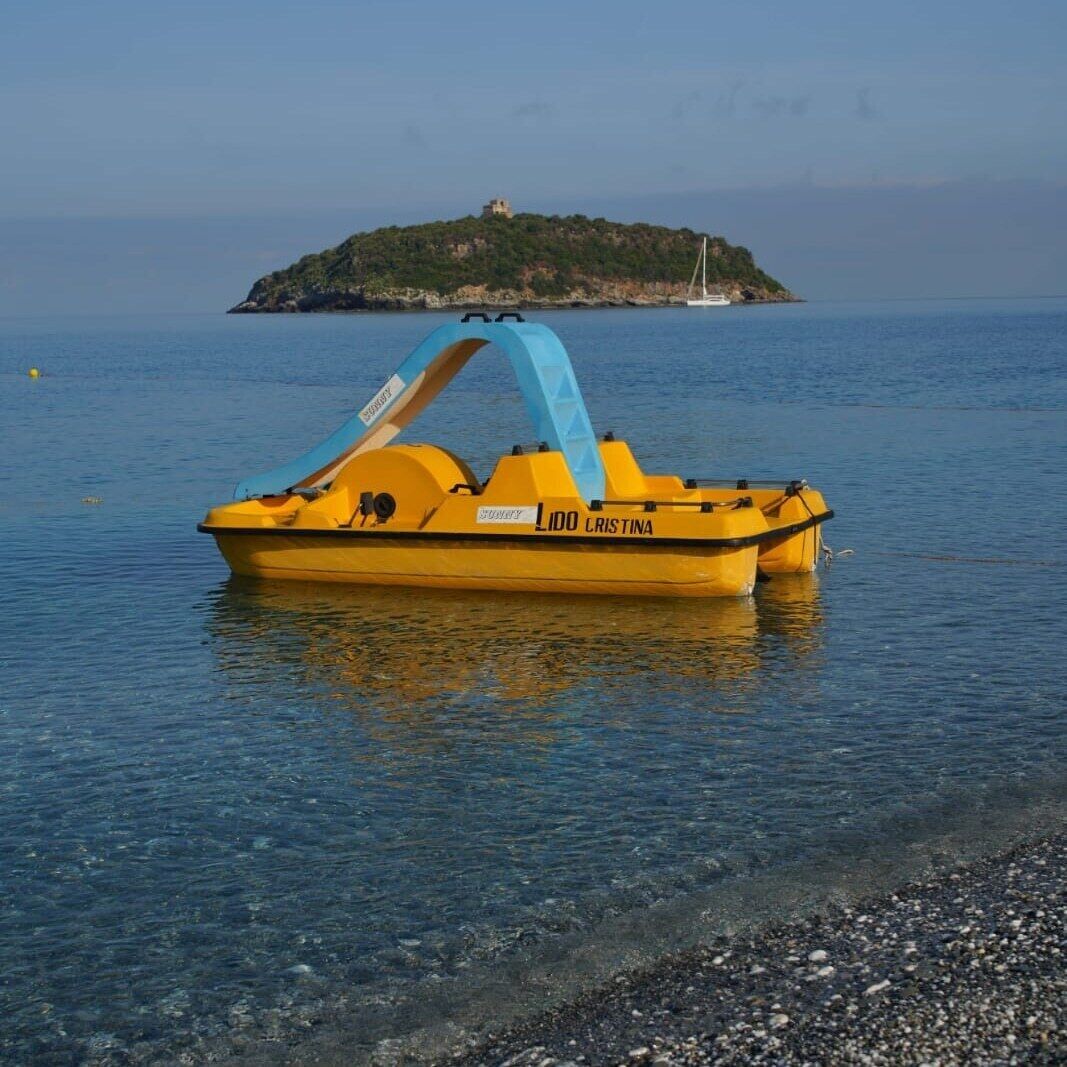 A yellow boat named "LIDO CRISTINA" docked at a pebble beach with clear water and a small island in the background.