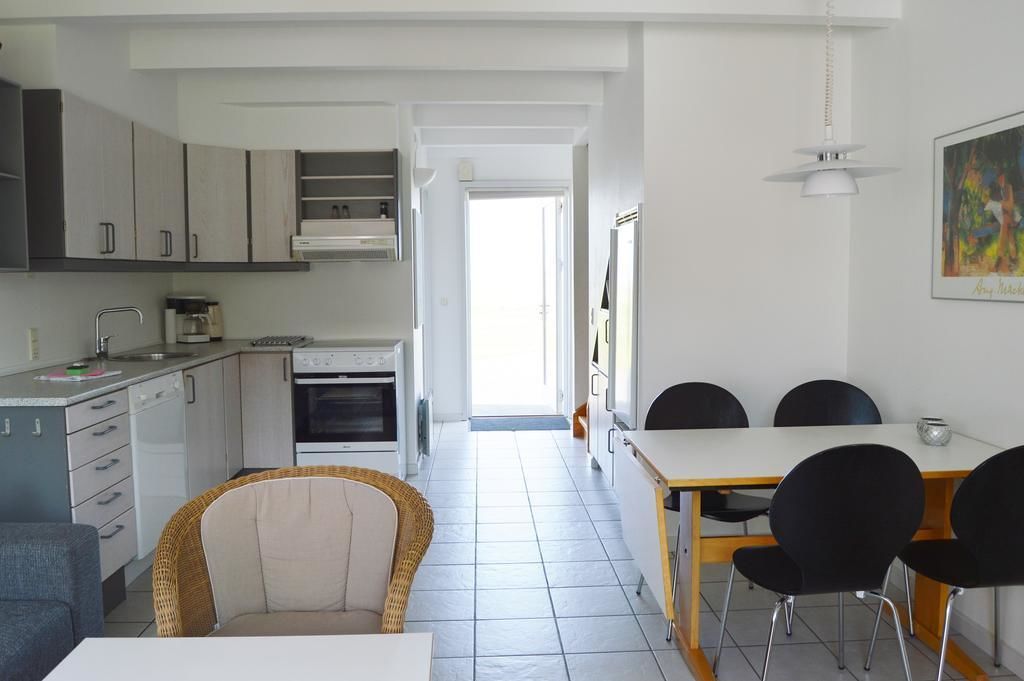 Ett modernt kök med vita skåp, en matplats med ett vitt bord och svarta stolar, och utsikt över gatan genom en öppen dörr.
