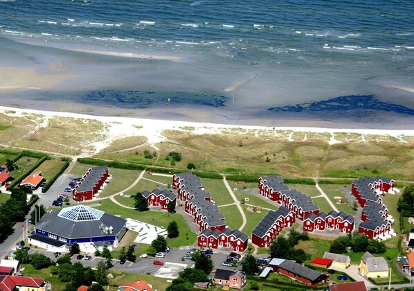 An aerial view of a coastal village with red houses, a large blue building with a dome, and a sandy beach.