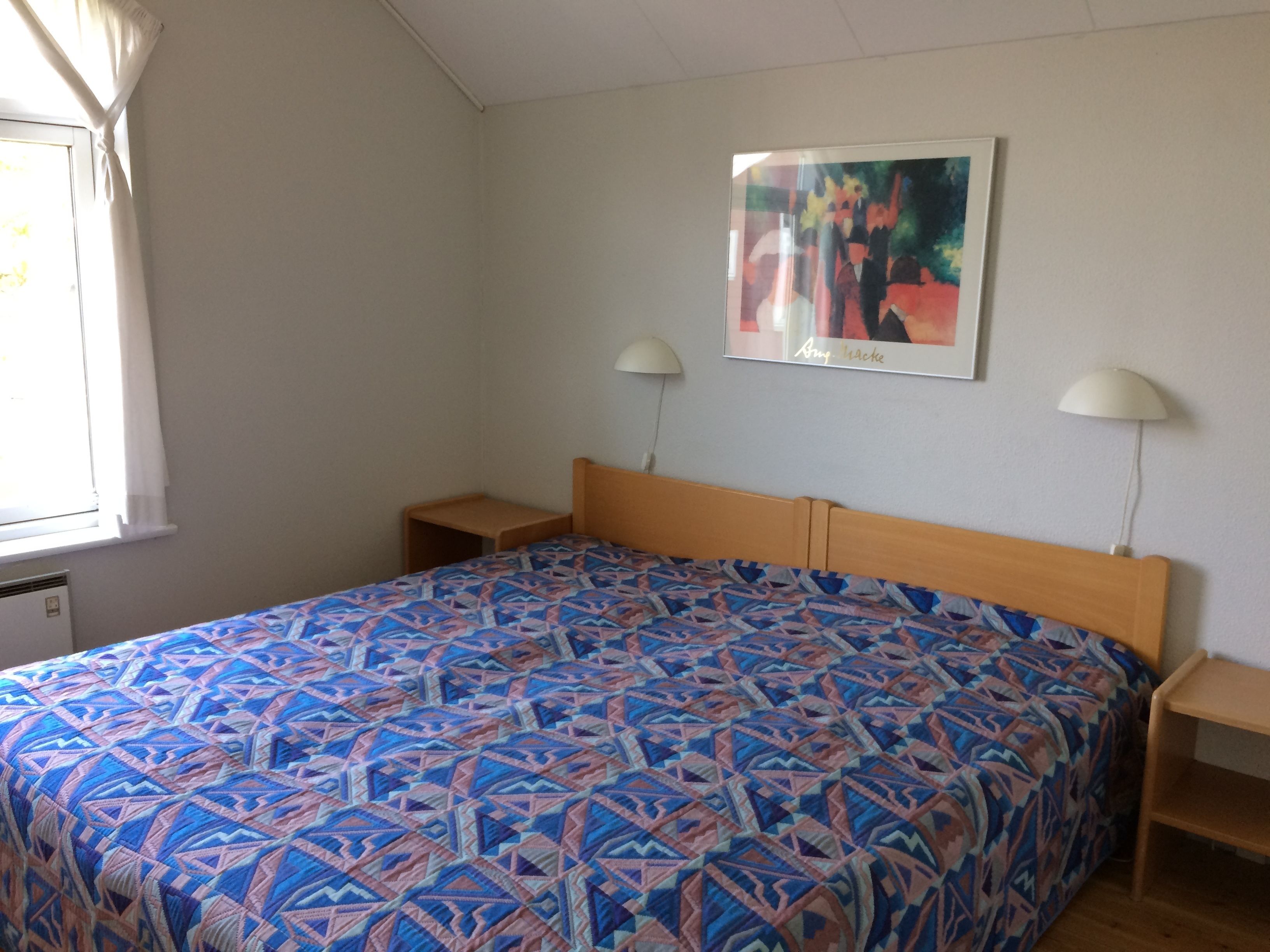 A bedroom with a patterned bedspread, two bedside tables, a wall-mounted lamp, and a framed picture.
