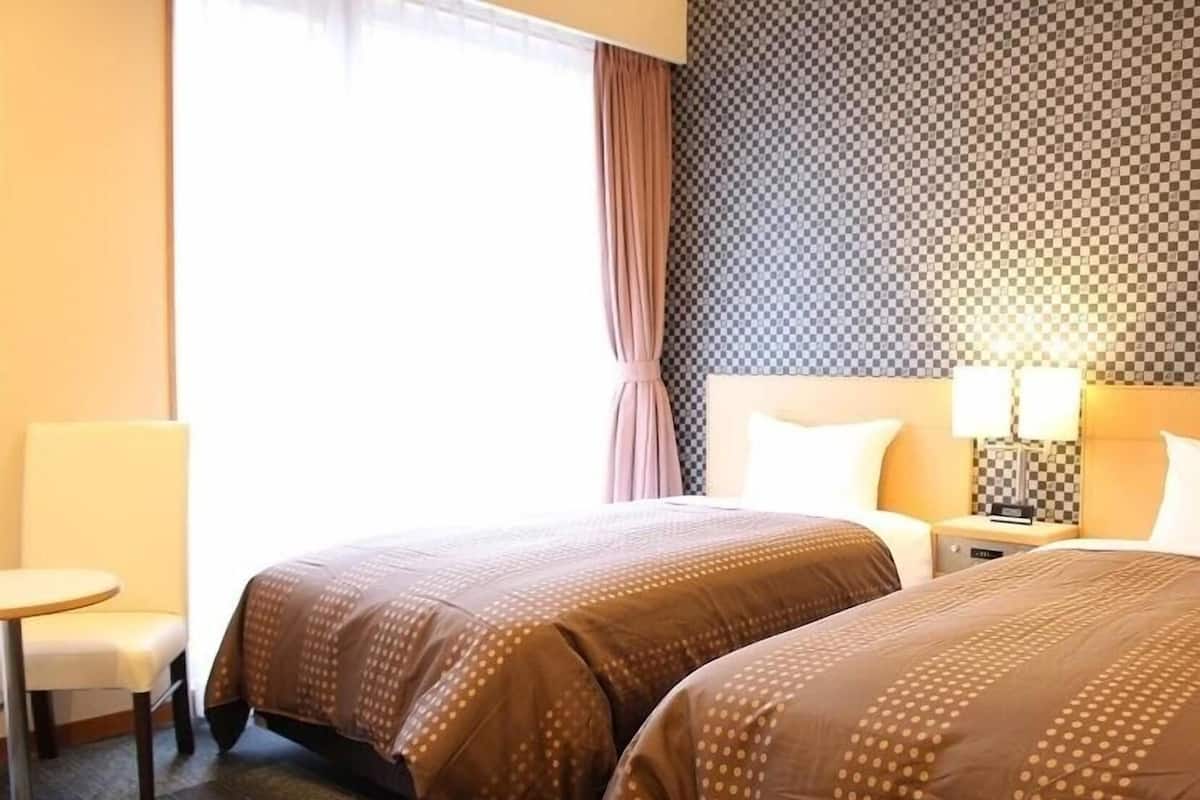 A hotel room with two beds, a chair, a nightstand with lamps, and a window with curtains.