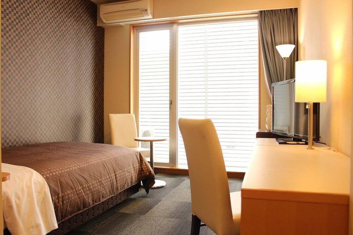 A hotel room with a bed, a desk, a chair, a TV, and a window with blinds.