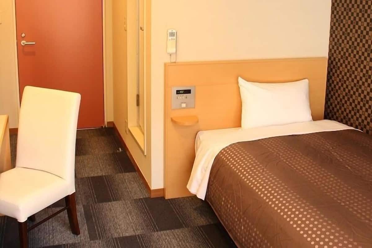 A hotel room with a bed, a chair, a wall-mounted phone, and a thermostat.