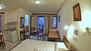 Habitación de hotel con cama, escritorio, silla, kitchenette pequeña y balcón con cortinas.