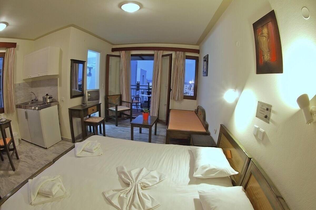 A hotel room with a bed, a desk, a chair, a small kitchenette, and a balcony with curtains.